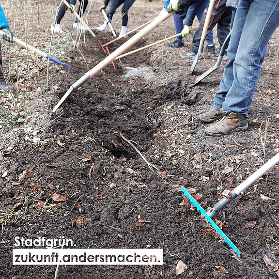 Menschen bei der Gartenarbeit in einem Waldgebiet, arbeiten mit Stechern und Schaufeln im Boden. Die Szene zeigt eine Gemeinschaftsaktion zur Verbesserung der Umwelt. Im Vordergrund steht der Slogan "Stadtgrün. Zukunft anders machen."