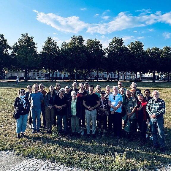 Rund 25 Erwachsene stehen dicht beieinander für ein Gruppenfoto auf einer Wiese. Im Hintergrund eine Baumallee. Spätes Sonnenlicht, klarer Himmel.