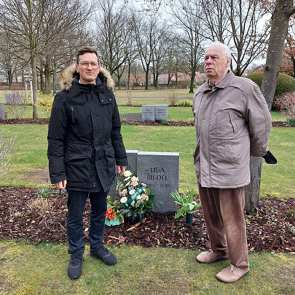 Zwei Männer stehen auf einem Friedhof neben einem geschmückten Grabstein mit Blumen. Sie blicken ernst; es ist ein ruhiger, winterlicher Tag. Die Szene vermittelt Gedenken an eine verstorbene Person.