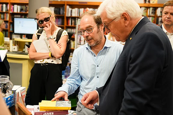 Eine Buchhandlungsszene, in der zwei Männer ein Buch betrachten. Einer von ihnen zeigt auf das Buch, während der andere interessiert zuhört. Im Hintergrund sind mehrere Personen zu sehen, die ebenfalls an der Buchhandlung interessiert sind.