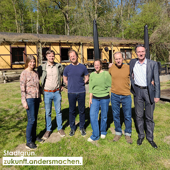 Sechs Personen stehen in einer Reihe im Freien vor einem gelben Gebäude. Die Gruppe besteht aus drei Männern und drei Frauen, die lächeln und sich näher zusammen positionieren. Im Hintergrund sind Bäume sichtbar. Der Schriftzug "Stadtgrün. zukunft.andersmachen." befindet sich am unteren Bildrand.