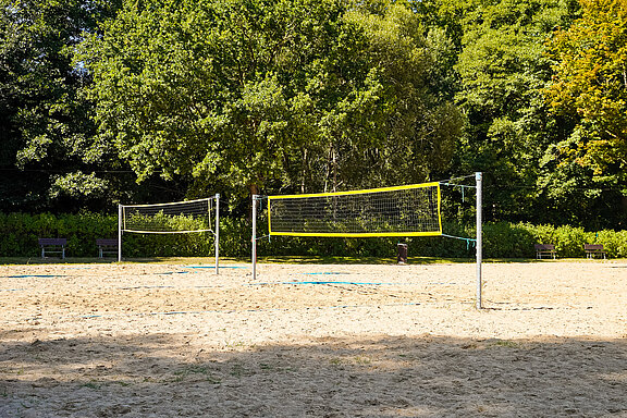 Zwei Volleyballnetze stehen auf einem Sandplatz, umgeben von Bäumen und Sträuchern. Im Hintergrund sind Bänke zu sehen, die zum Verweilen einladen.