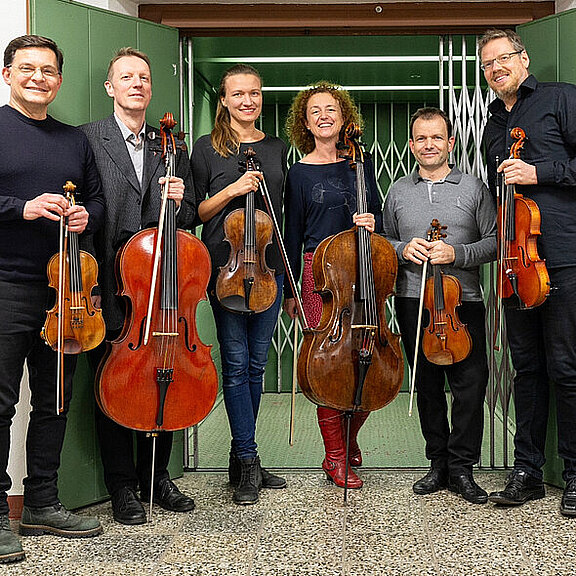 Streichsextett des Brandenburgischen Staatsorchesters um Stefan Hunger (Fotoautor Peter Dynow) Sechs Musiker stehen in einer Reihe vor einer offenen Tür, jeder hält ein Streichinstrument. Die Gruppe besteht aus drei Männern und drei Frauen, die zwischen Geigen und Bratschen variieren. Sie lächeln und wirken entspannt. Hinter ihnen ist ein grüner Hintergrund sichtbar.