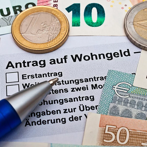 Formular „Antrag auf Wohngeld“ mit Auswahlkästchen, daneben Euro-Banknoten und -Münzen sowie ein Kugelschreiber – Symbol für Antragstellung staatlicher Wohnkostenhilfe.