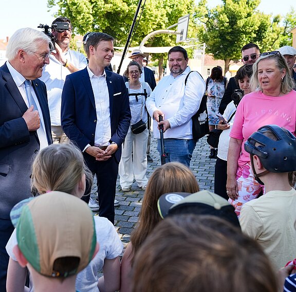 Eine Gruppe von Menschen, darunter zwei Männer in Anzügen und eine Frau in einem pinken T-Shirt, steht in einem Freiluftbereich. Sie sprechen mit Kindern, die Hüte tragen. Im Hintergrund sind weitere Erwachsene und grüne Bäume zu sehen.