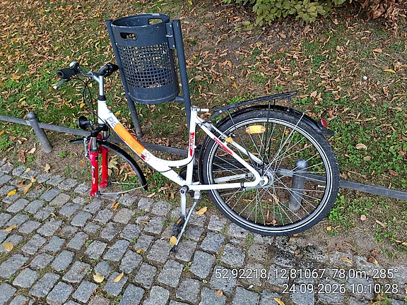 Ein weiß-rotfarbenes Fahrrad ohne Vorderrad steht schräg an einem kurzen Poller auf einem gepflasterten Weg. Daneben eine Grasfläche mit Herbstlaub.