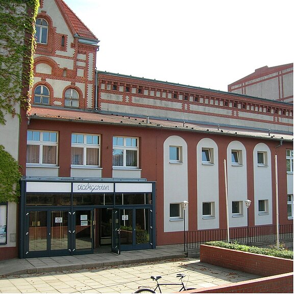 Historisches Backsteingebäude mit dekorierter Fassade. Im Vordergrund ein moderner Eingangsbereich mit Glasfront und Vordach. Reihe von rundbogigen Fenstern und höherer Gebäudeteil dahinter. Fußweg und niedrige Mauer davor.