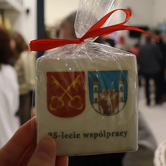Hand hält ein in Folie verpacktes, quadratisches Souvenir/Keks mit rotem Band. Vorderseite zeigt zwei Wappen und die polnische Aufschrift „25‑lecie współpracy“ (25 Jahre Zusammenarbeit).