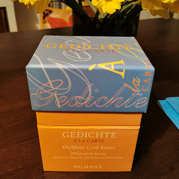 Eine Produktbox mit dem Titel "GEDICHTE A LA CARTE" in blauer und orangefarbener Gestaltung. Die Box enthält 200 poetische Karten mit dem Thema Leben, Sinne, Versöhnung und Verwobenheit, herausgegeben von Dumont. Im Hintergrund sind gelbe Blumen sichtbar.