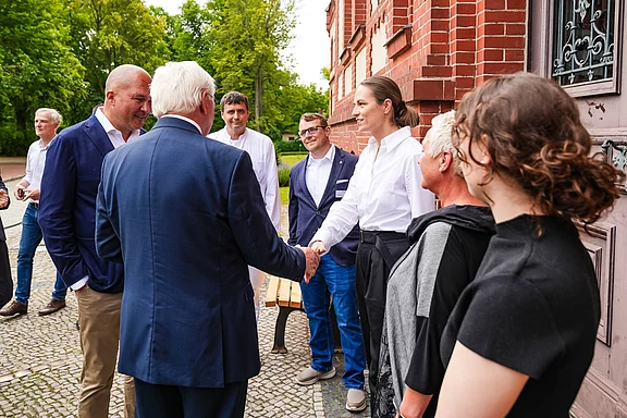 Eine Gruppe von Personen steht im Freien vor einem historischen Gebäude. Eine Frau in einem weißen Hemd schüttelt einem Mann die Hand. Die anderen Anwesenden beobachten die Begegnung und lächeln, während Bäume im Hintergrund sichtbar sind.