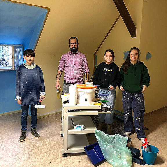 Eine Gruppe von vier Personen steht in einem unrenovierten Raum. Sie sind mit Malerwerkzeug ausgestattet; einer hält eine Farbrolle. Im Hintergrund sind Farbbehälter und Utensilien auf einem Tisch sichtbar, während die Wände teilweise in Hellblau gestrichen sind.