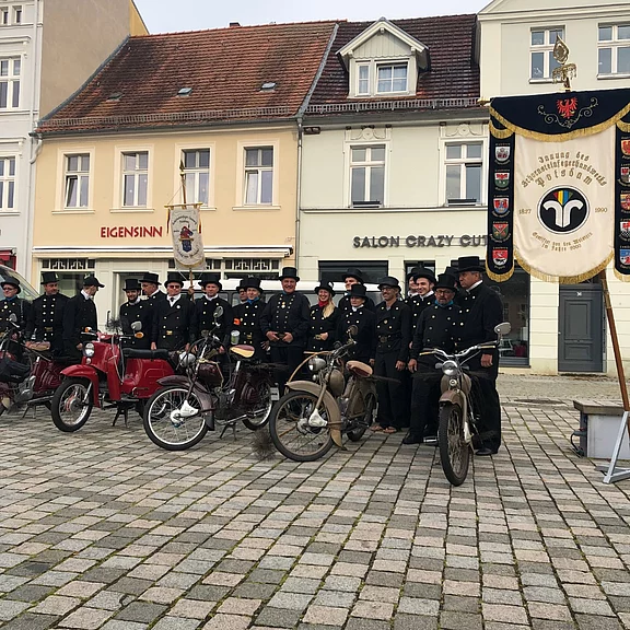 Eine Gruppe von Männern in Uniformen steht in einer historischen Stadtmitte vor mehreren Motorrädern. Im Hintergrund sind alte Gebäude und ein Banner zu sehen. Die Szene vermittelt ein Gefühl von Tradition und Gemeinschaft bei einem besonderen Anlass.