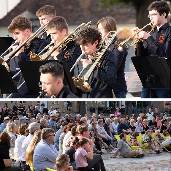 Oben spielen junge Bläser in schwarzen Hemden Trompeten und Posaunen an Notenständern. Unten sitzt eine große Menschenmenge im Freien und hört zu. Szene eines Open-Air-Konzerts auf einem Platz.