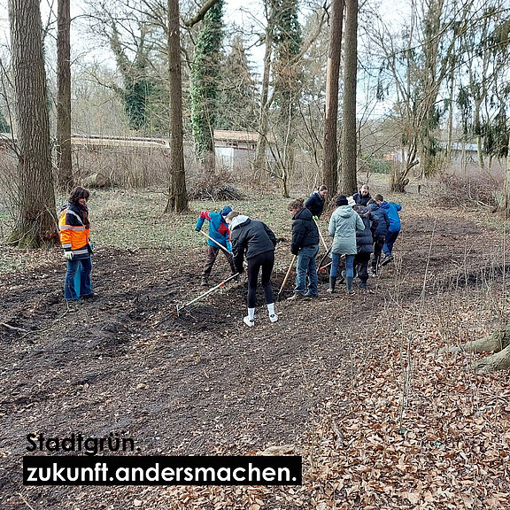 Eine Gruppe von Menschen arbeitet in einem Waldgebiet. Sie graben in der Erde und pflanzen Bäume oder Sträucher. Im Hintergrund sind hohe Bäume zu sehen. Ein Banner mit dem Text „StadtGrün: zukunft.andersmachen“ ist sichtbar.