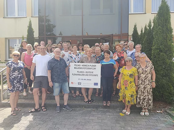 Gruppenfoto vor einem Gebäude: Viele Erwachsene, überwiegend ältere, lächeln in Sommerkleidung. In der Mitte halten Teilnehmende ein großes Schild mit der Aufschrift zu einem polnisch-deutschen Pleinair für Malerei und Fotografie, mit Datum 17.–19.06.2022. Gemeinschaftliches, freundliches Event.