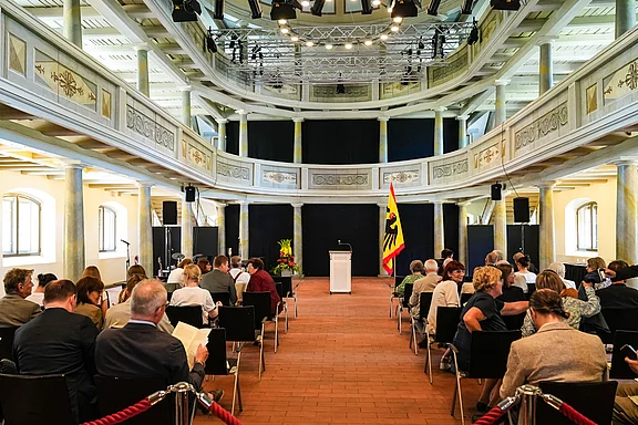 Ein geschmackvoll dekorierter Veranstaltungsraum mit Sitzreihen, die auf ein Rednerpult ausgerichtet sind. Im Hintergrund steht eine Landesflagge. Die Besucher, in verschiedenen Kleidungsstilen, warten auf den Beginn der Veranstaltung. Die Atmosphäre scheint formal und erwartungsvoll zu sein.