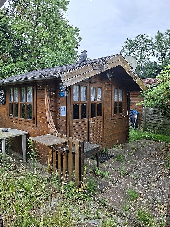 Kleines hölzernes Gartenhaus mit Fensterfront und Tür, umgeben von überwuchertem Grün. Auf dem Satteldach sitzt eine Taube. Vor dem Haus liegen verwitterte Pflastersteine und ein niedriges Holztor.