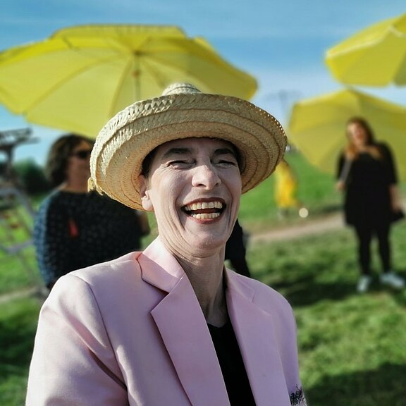 Lachende Person mit Strohhut und rosa Blazer im Vordergrund, bei Sonnenschein im Freien. Im Hintergrund unscharf gelbe Sonnenschirme und wenige weitere Menschen. Fröhliche, sommerliche Stimmung.