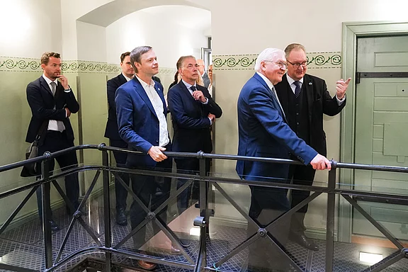 Eine Gruppe von Männern in formeller Kleidung steht auf einer Treppe und hört einem älteren Mann zu, der spricht und gestikuliert. Sie befinden sich in einem hellen Innenraum mit historischen Elementen.