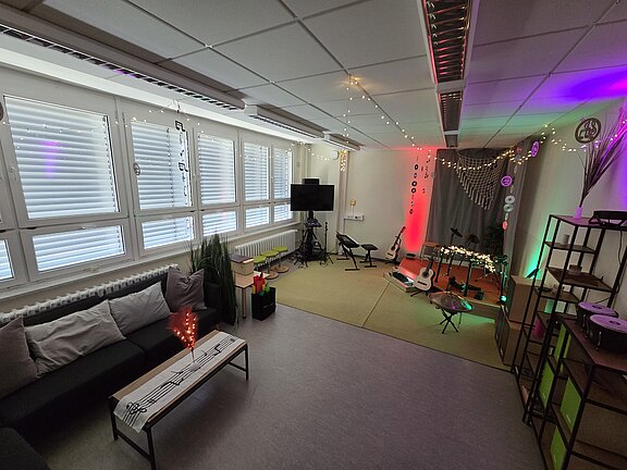 Kleiner Proberaum bzw. Wohnzimmer-Bühne mit farbiger Beleuchtung. Im hinteren Bereich stehen Mikrofonständer, Gitarren und Hocker vor einem Vorhang; vorn eine Sitzecke mit Sofa und Couchtisch. Kabel, Notenständer und Monitor bereiten eine intime Musikaufführung vor.