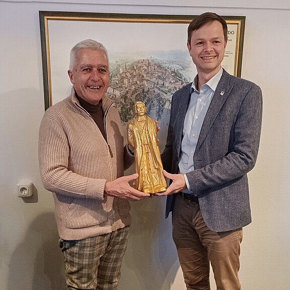 Zwei Männer stehen nebeneinander, lächelnd, und halten eine goldene Statue einer Person. Der Mann links trägt einen beigen Pullover und der rechts einen blauen Blazer. Im Hintergrund ist ein dargestelltes Stadtpanorama zu sehen.