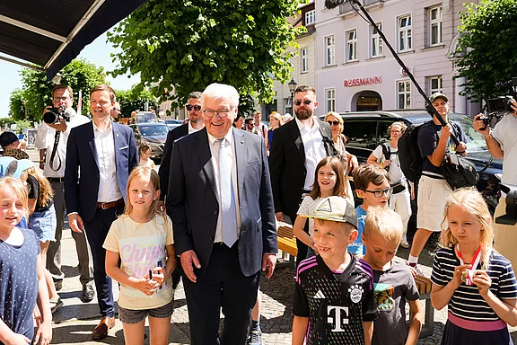 Eine Gruppe von Kindern steht auf der Straße und lächelt, während ein älterer Mann in Anzug und Brille, umgeben von Begleitpersonen, an ihnen vorbeigeht. Einige Erwachsene filmen die Szene mit Kameras. Die Atmosphäre ist fröhlich und unbeschwert.