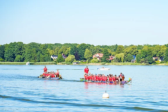 Zwei Drachenboote mit Ruderern in roten Outfits fahren auf einem ruhigen See. Im Hintergrund sind grüne Bäume und einige Häuser zu sehen, während die Sonne scheint. Die Atmosphäre wirkt sportlich und fröhlich.