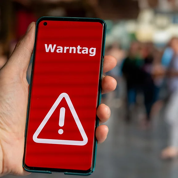Eine Hand hält ein Smartphone, auf dem der Text „Warntag“ auf rotem Hintergrund und ein Warnsymbol in Form eines Ausrufezeichens in einem Dreieck zu sehen ist.