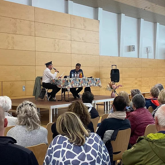 Vortragsabend in einer Halle: Zwei Personen sitzen an einem Tisch auf einer Bühne; eine Person mit Mütze spricht ins Mikrofon, die andere liest. Vor der Bühne sitzt ein größeres Publikum und hört zu.