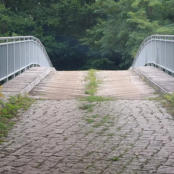 Verlassene Fußgängerbrücke im Grünen: Steinweg führt auf eine hölzerne Brücke mit Metallgeländern, deren Mitte stark abgesackt und von Gras durchwachsen ist – wirkt beschädigt und unbenutzbar.