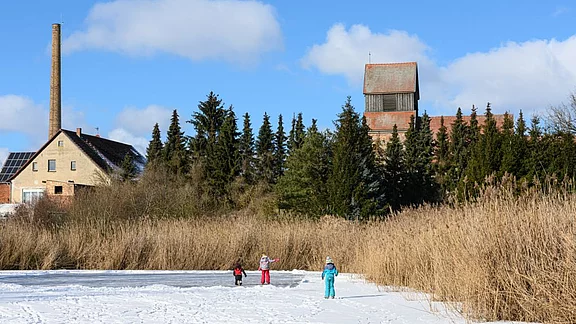 Im Vordergrund spielen Kinder auf verschneitem Boden. Im Hintergrund sind ein Gebäude mit Schornstein und eine Kirche sichtbar, umgeben von Bäumen und hohem Gras. Der Himmel ist blau mit wenigen Wolken.