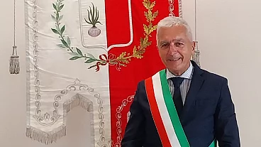 Älterer Mann im dunklen Anzug trägt eine Schärpe in den italienischen Farben. Er steht lächelnd vor einem rot-weißen Gemeindebanner mit Wappen und der Aufschrift „Comune di Certaldo“.