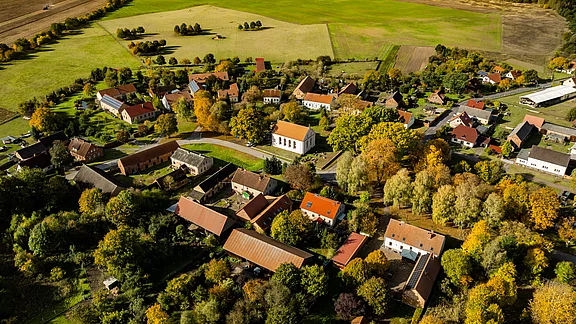 Eine Luftaufnahme eines kleinen dorfs mit roten Dächern, umgeben von grünen Feldern und Wäldern. Die Bäume zeigen Herbstfarben. Die Straßen sind klar sichtbar, und einige Gebäude sind gruppiert. Insgesamt vermittelt das Bild eine ruhige, ländliche Atmosphäre.