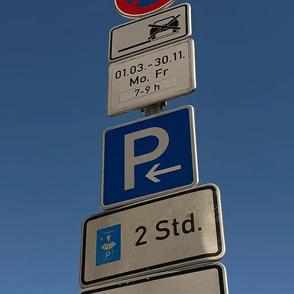 Verkehrsschild: Blaues P mit Pfeil nach links. Darunter: Parken mit Parkscheibe, maximal 2 Stunden. Zusatzschilder oben mit zeitlichen Einschränkungen (März bis November, Mo–Fr, 7–9 Uhr).