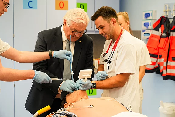 Eine Gruppe von medizinischem Personal und einem Politiker führt eine medizinische Simulation durch. Sie arbeiten zusammen an einem Patientensimulationsmodell, wobei sie medizinische Geräte und Techniken verwenden. Die Szene zeigt Teamarbeit in einer Ausbildungssituation im Gesundheitswesen.