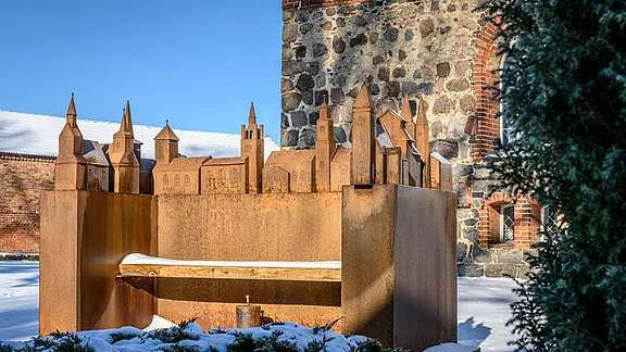 Die Abbildung zeigt eine kunstvolle Metallskulptur, die eine historisch inspirierte Stadtansicht darstellt. Darin sind verschiedene Türme und Gebäude zu erkennen. Im Vordergrund befindet sich eine Bank auf einer schneebedeckten Fläche, umgeben von grünen Sträuchern.