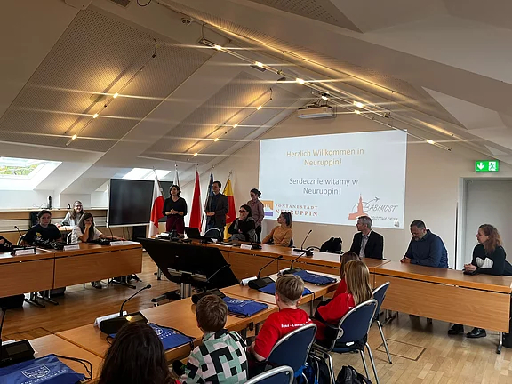 Konferenzraum mit U-förmigen Tischen und Mikrofonen. Eine Person spricht vor einer Projektionswand mit der Folie Herzlich Willkommen in Neuruppin – Serdecznie witamy w Neuruppin. Teilnehmende, darunter Jugendliche und Erwachsene, sitzen rundum; Nationalflaggen im Hintergrund deuten auf ein internationales Treffen hin.
