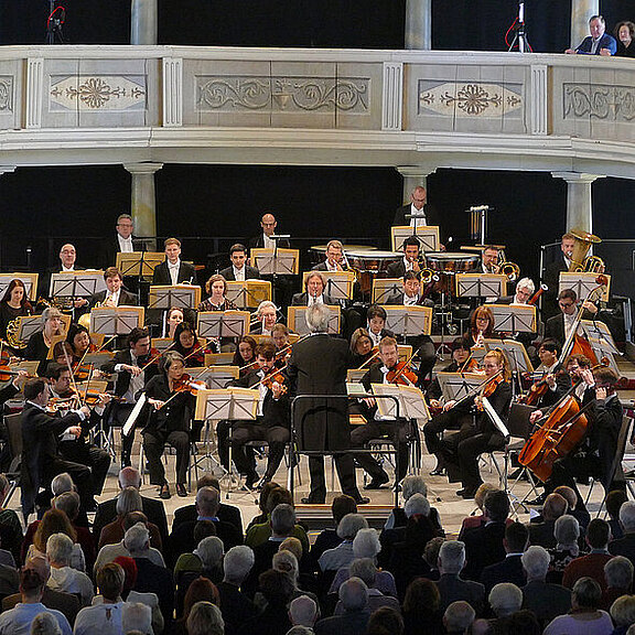 Ein Orchester tritt auf einer Bühne auf. Der Dirigent steht vor den Musikern, die verschiedene Instrumente spielen. Im Publikum sind zahlreiche Zuhörer sichtbar, die auf die Darbietung achten. Die Atmosphäre ist feierlich und konzentriert.