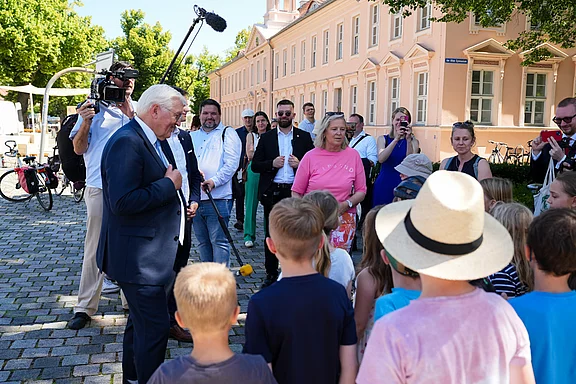 Eine Gruppe von Kindern steht um einen Mann in Anzug, der mit ihnen spricht. Im Hintergrund sind Erwachsene und Kamerateams zu sehen. Die Szene findet draußen an einem sonnigen Tag statt, vor einer hellen Gebäudefassade.