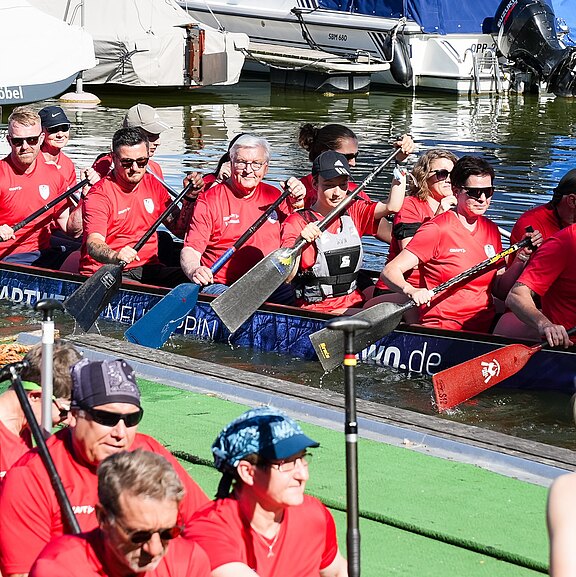 Eine Gruppe von Sportlern in roten T-Shirts sitzt in einem Ruderkahn und bereitet sich auf das Rudern vor. Die Umgebung zeigt Wasser und Boote im Hintergrund. Das Team ist auf den Sport konzentriert und hält Paddel bereit.