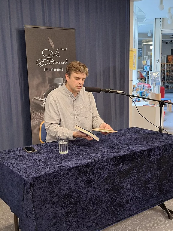 Ein Mann sitzt an einem Tisch mit dunklem Tuch und liest aus einem aufgeschlagenen Buch in ein Mikrofon. Vor ihm stehen ein Glas Wasser und ein Handy. Die Szene wirkt wie eine öffentliche Lesung oder Literaturpreis-Veranstaltung.