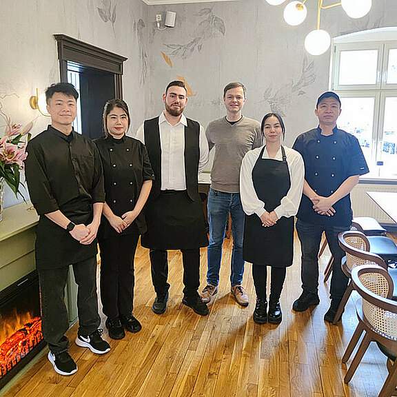 Sechs Personen stehen in einem Restaurant. Alle tragen Küchen- oder Servicekleidung, während sie lächeln. Im Hintergrund ist eine stilvolle Wandgestaltung und Möbel zu sehen. Die Atmosphäre wirkt einladend und freundlich.