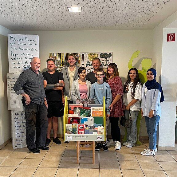 Eine Gruppe von acht Personen steht in einem Raum. Im Vordergrund ist ein Bücherregal mit verschiedenen Büchern und Materialien sichtbar. An der Wand hängen Kunstwerke, und es gibt eine informative Tafel. Die Atmosphäre wirkt freundlich und einladend.