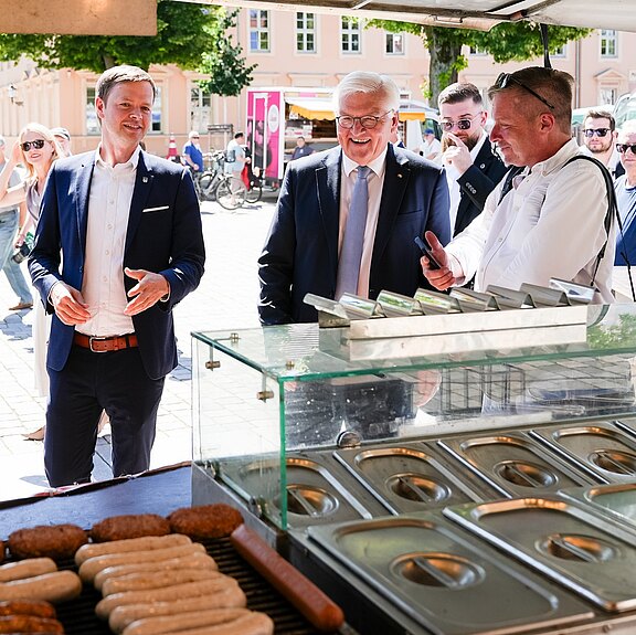 Eine Gruppe von Personen steht an einem Verkaufsstand im Freien. Ein Mann in Anzug spricht mit einem lächelnden älteren Mann, während ein weiterer Mann im Hintergrund zuschaut. Auf dem Stand sind verschiedene Grillgerichte sichtbar, darunter Bratwürste und Frikadellen.