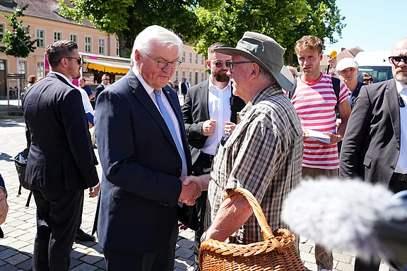 Der Bundespräsident shake hands with einem älteren Mann, der einen Korb hält. Im Hintergrund stehen mehrere Menschen in einem urbanen Umfeld. Es herrscht eine freundliche Atmosphäre.