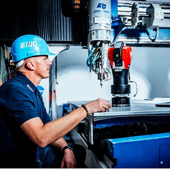 Ein Industriearbeiter mit blauem Schutzhelm beobachtet einen roten kollaborativen Roboter, der auf einer Werkbank an einem Metallteil arbeitet; moderne Maschinen im Hintergrund, Szene zeigt Mensch‑Roboter‑Zusammenarbeit in der Fertigung.