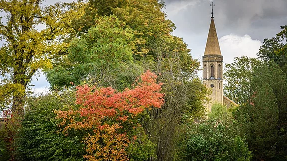 Eine farbenfrohe Landschaft im Herbst mit bunten Bäumen, darunter ein strahlend rotes Laub. Im Hintergrund ragt eine Kirche mit spitzem Turm in die Höhe, umgeben von grünen Sträuchern und Bäumen. Der Himmel ist bewölkt und verleiht der Szene eine ruhige Atmosphäre.