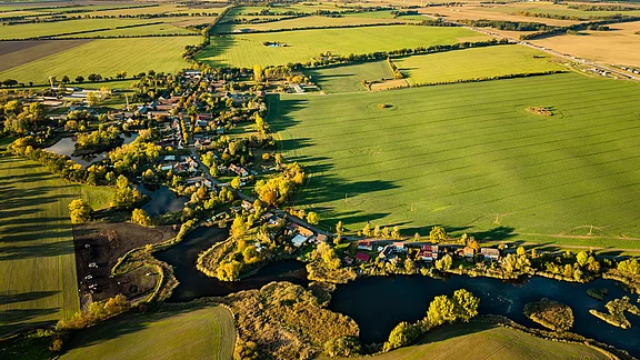 Luftaufnahme eines ländlichen Gebiets mit einem kleinen Dorf, umgeben von grünen Feldern und Bäumen. Ein Fluss schlängelt sich durch die Landschaft, während im Hintergrund Windkraftanlagen sichtbar sind. Die Szenerie vermittelt eine friedliche, natürliche Umgebung.
