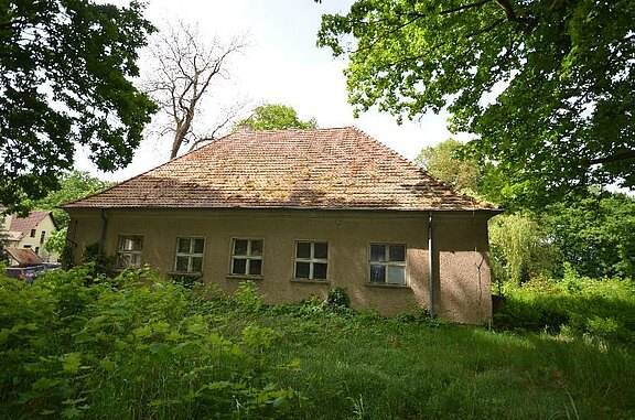 Ansicht von der Straße Ein verfallenes, einst bewohntes Haus mit grauer Fassade und einem schrägen, mit Moos bedeckten Dach. Umgeben von hohem Gras und Bäumen, strahlt es eine verlassene und ruhige Atmosphäre aus. Die Fenster sind teilweise verschmutzt und geben den Eindruck von Vergänglichkeit.