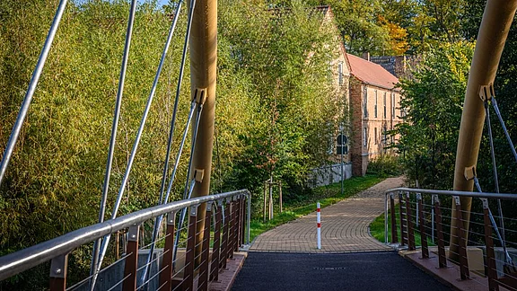 Blick auf eine schmale Fußgängerbrücke, die über einen Weg führt. Im Hintergrund ist ein Gebäude mit rotem Dach, umgeben von üppigem Grün. Der Weg verläuft sanft zwischen Bäumen und Sträuchern, was eine ruhige, naturnahe Atmosphäre schafft.